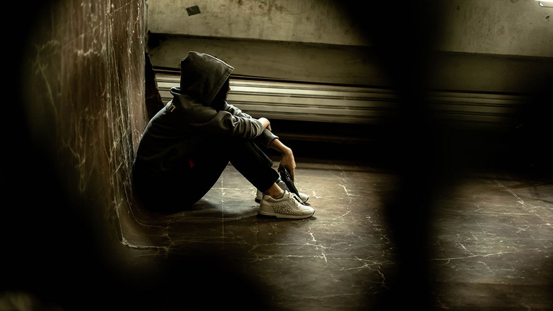 A person wearing a hoodie and sneakers is sitting on the floor with knees bent and arms resting on the knees, in a dimly lit room with a radiator against the wall.